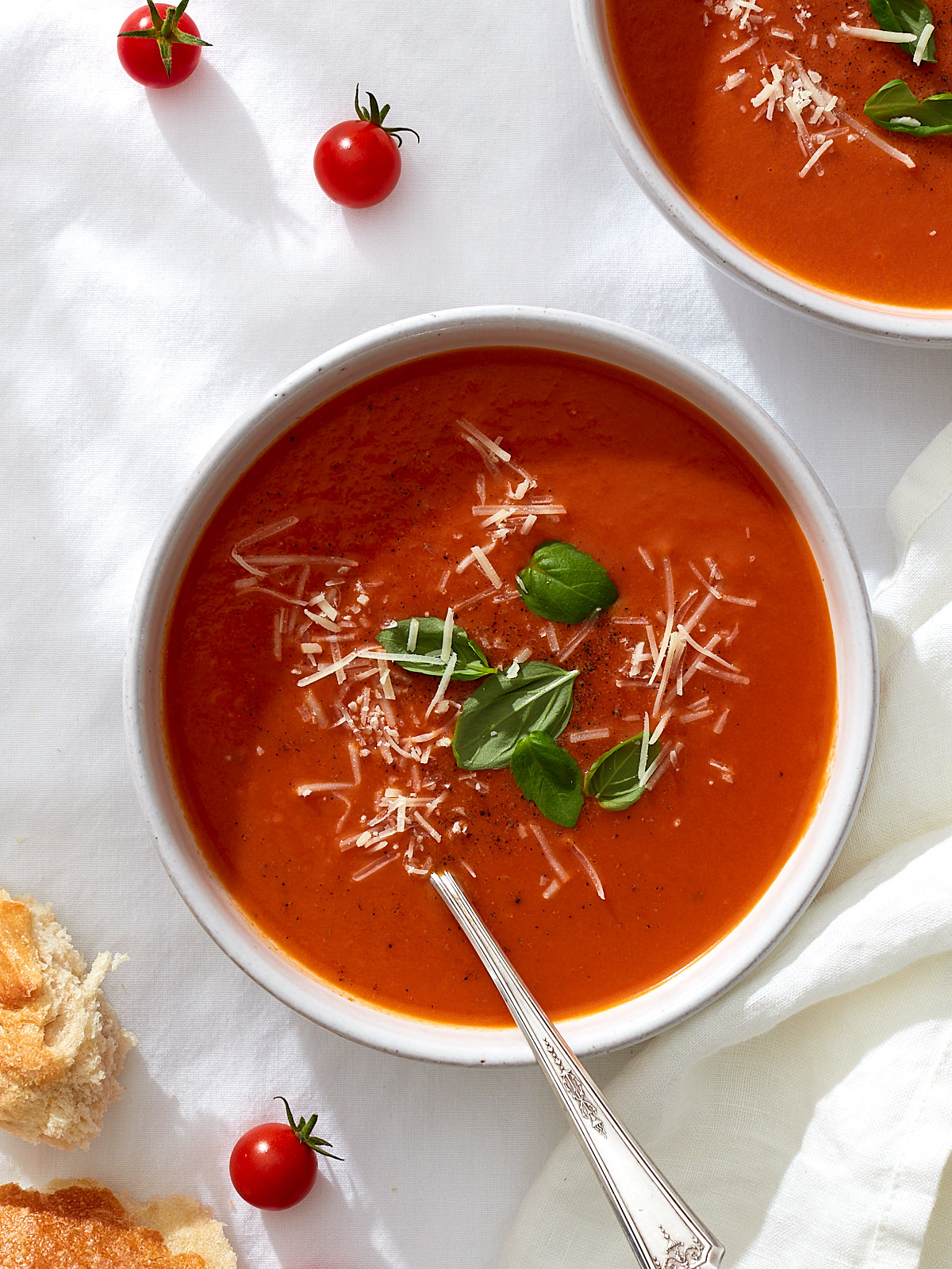 Two bowls of homemade roasted tomato soup with cracked black pepper, fresh basil leaves, and grated parmesan on a white linen table.