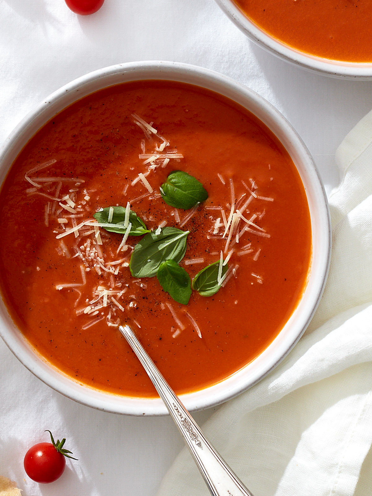 A bowl of homemade roasted tomato soup with cracked black pepper, fresh basil leaves, and grated parmesan on a white linen table.