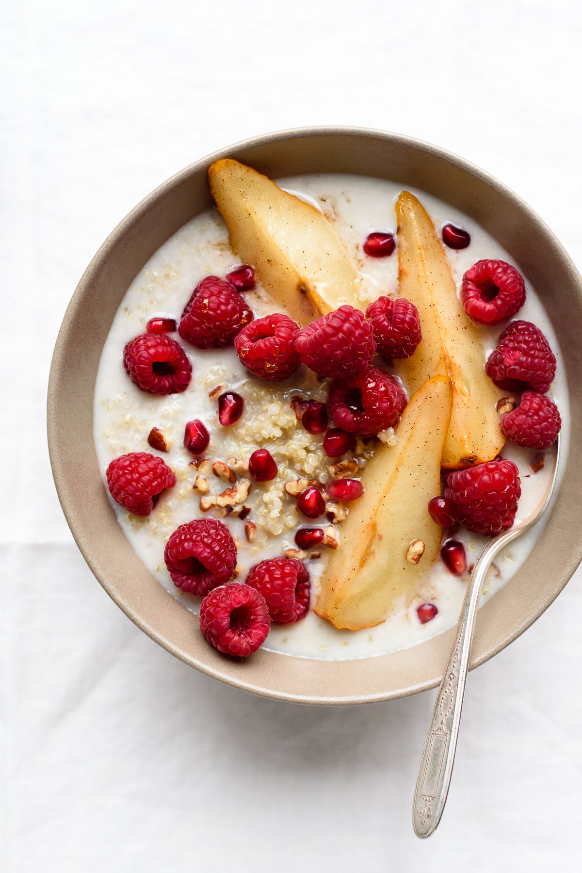 A bowl of quinoa breakfast porridge with roasted pears, fresh raspberries, pomegranate, pecans, and almond milk on a white linen tablecloth.