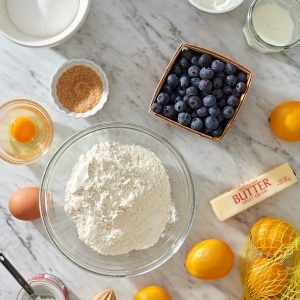 Baking ingredients (flour, sugar, eggs, sour cream, butter, milk, blueberries, salt, baking powder, lemons, and vanilla) on a marble background, ready to made into blueberry muffins.