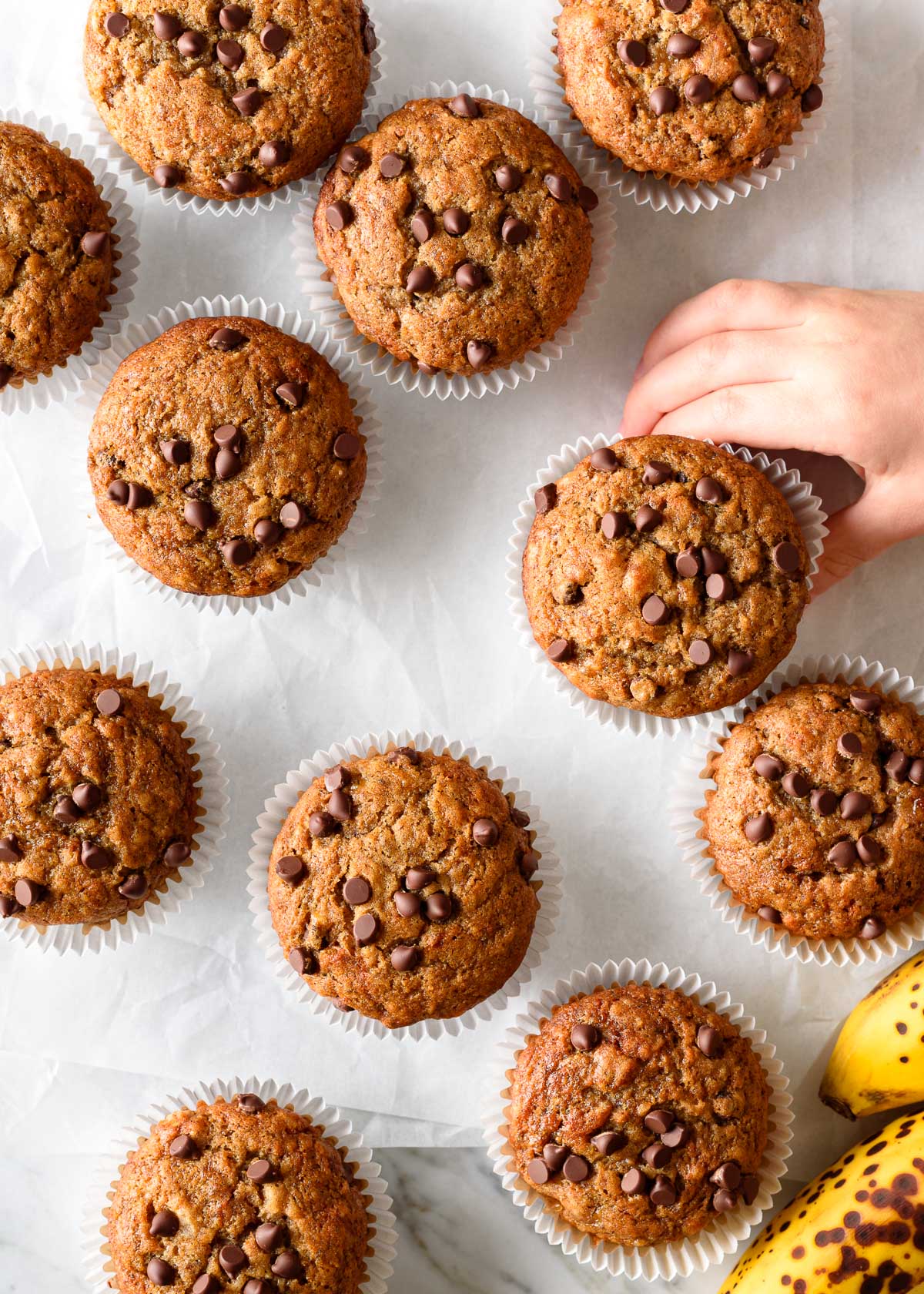 A toddler's hand reaching for a chocolate chip banana muffin on a white parchment paper background.