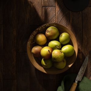 A rustic bowl of small Seckel pears on a dark wood table in strong shadow.