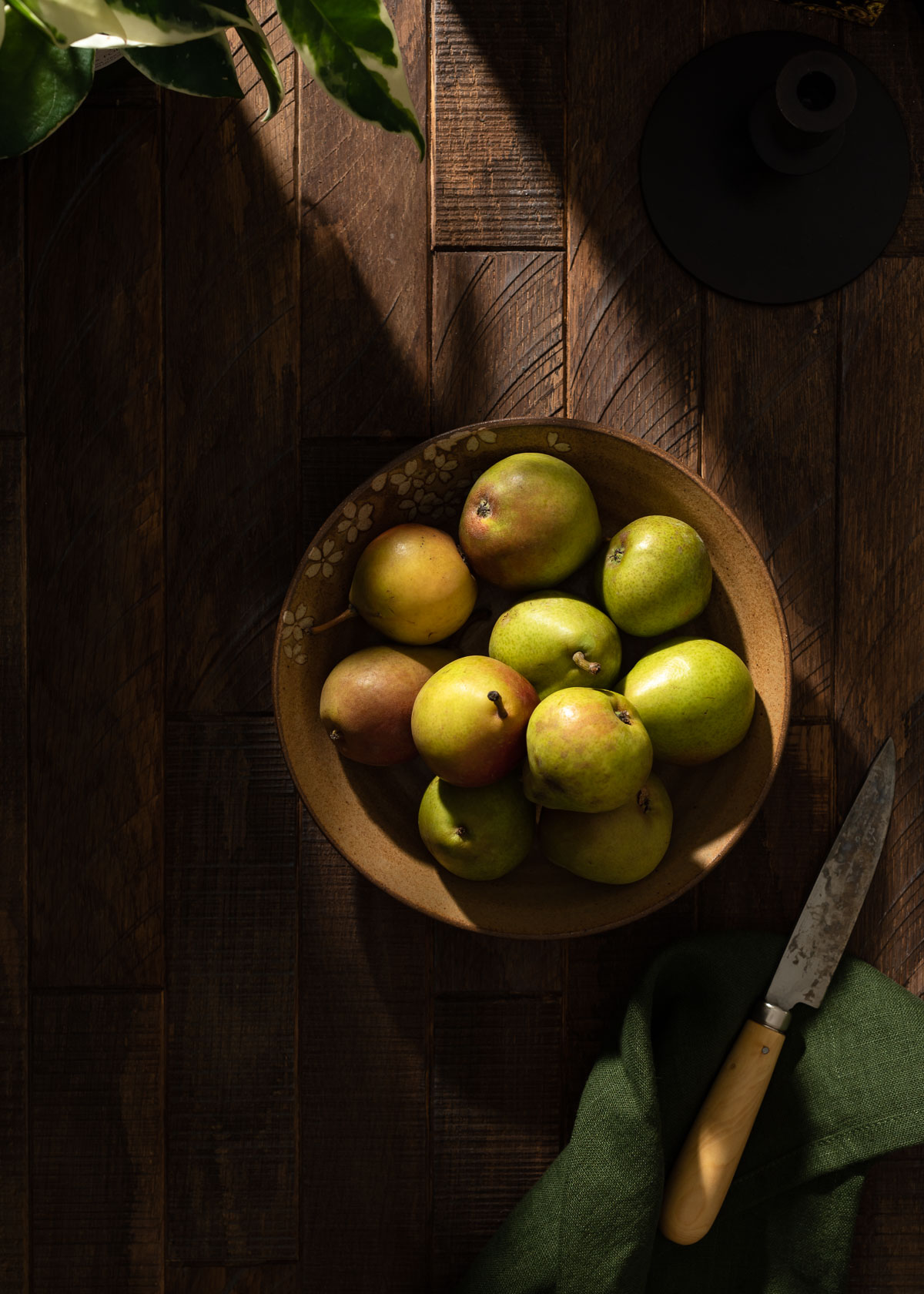 A rustic bowl of small Seckel pears on a dark wood table in strong shadow.