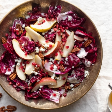 A large salad of radicchio, fresh pears, goat cheese, pecans, and pomegranate in a gold bowl on a linen tablecloth.