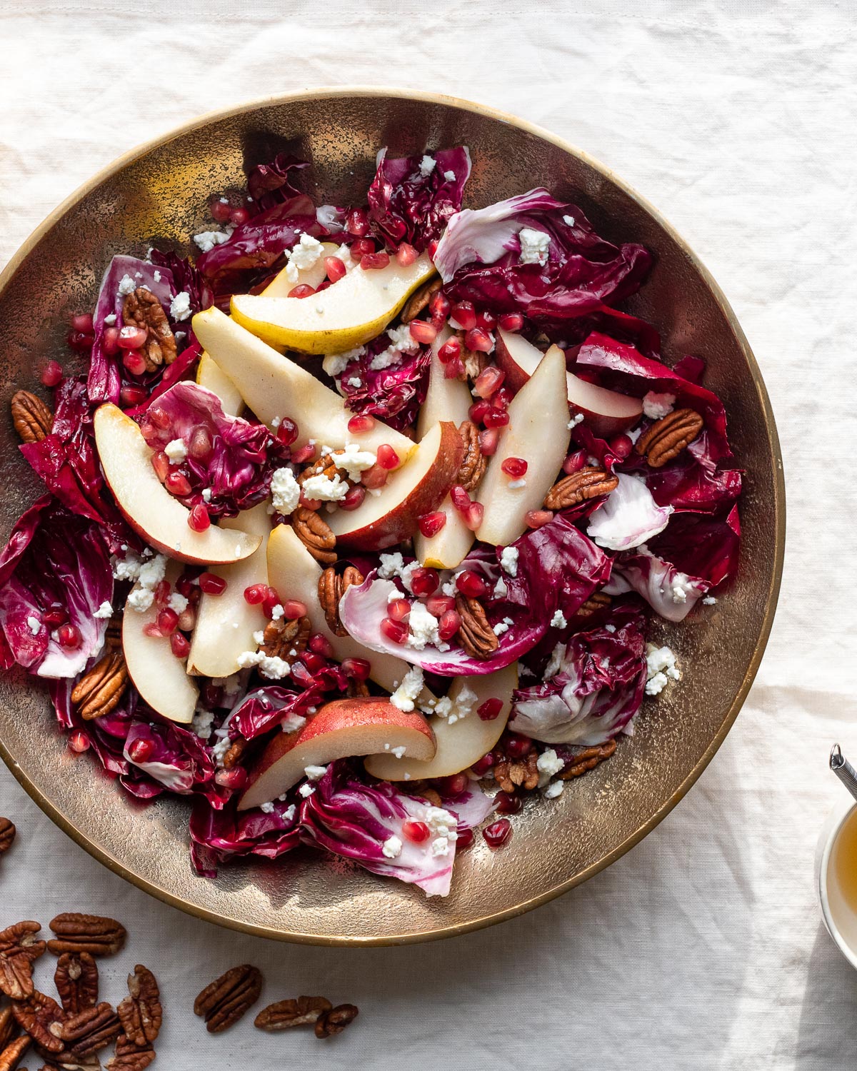 A large salad of radicchio, fresh pears, goat cheese, pecans, and pomegranate in a gold bowl on a linen tablecloth.