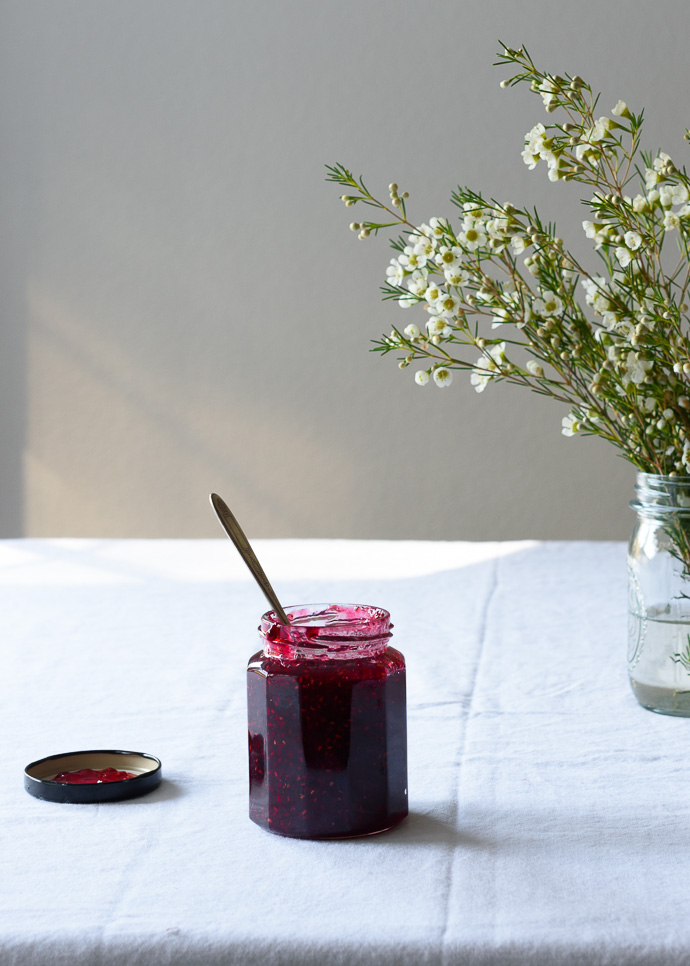 a jar of raspberry jam on a table with a vase of flowers behind.