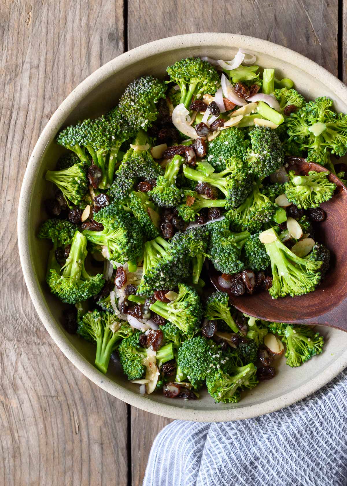 A bowl of broccoli salad with shallots, almonds, raisins, and a lemon tahini dressing.