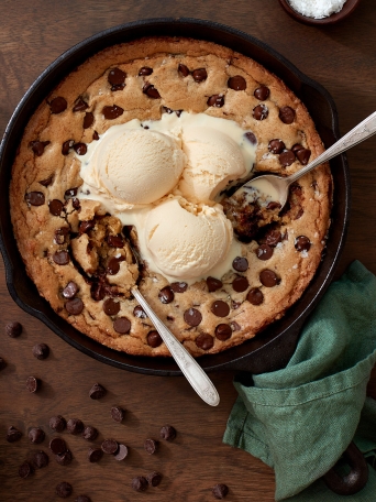 A warm from the oven chocolate chip skillet cookie topped with vanilla ice cream on a wood table.