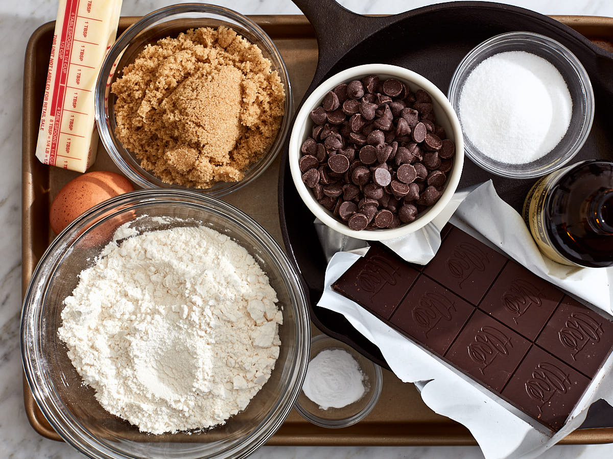 The ingredients to make a skillet chocolate chip cookie (flour, sugar, brown sugar, butter, egg, baking powder, salt, chocolate chips and a chocolate bar) in prep bowls on a baking sheet.