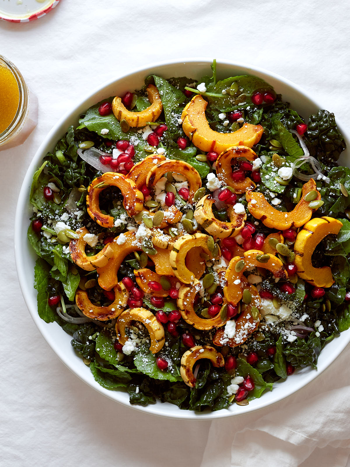 A roasted delicata squash and kale salad with goat cheese, pomegranate, pepitas, and shallots in a large white bowl on a white linen background.