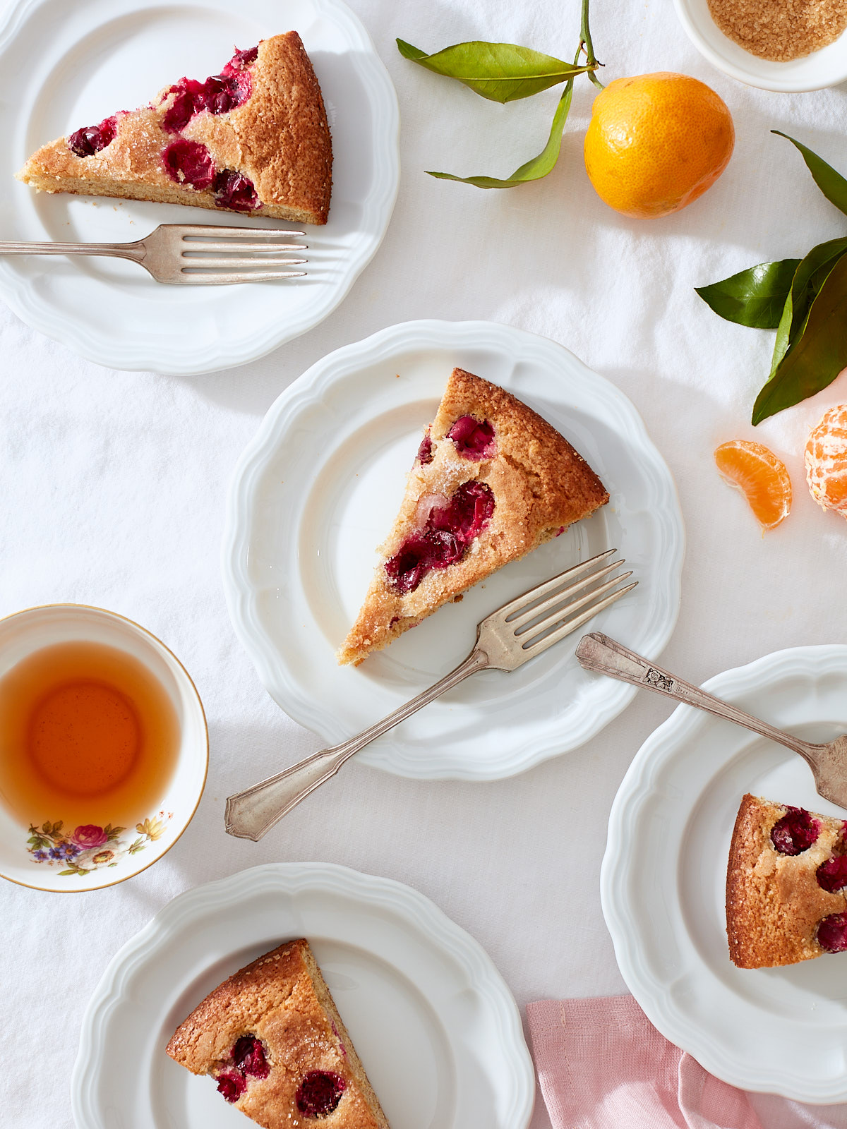 Citrus and Cranberry Buttermilk Cake | Slices of cranberry cake on white porcelain dessert plates with vintage forks, pink linen, tea, and fresh tangerines.
