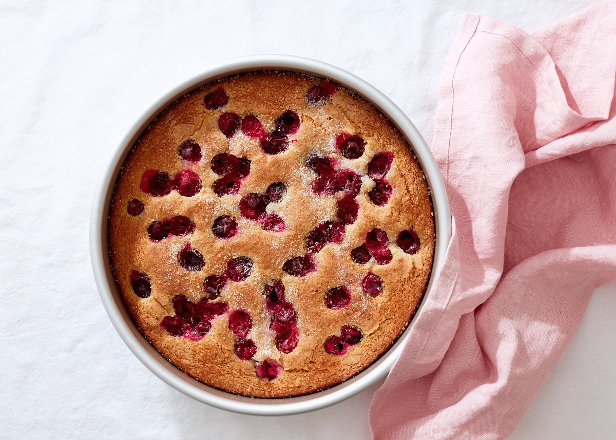 A simple cranberry cake warm from the oven in a round cake pan with a pink linen towel on a white linen table.