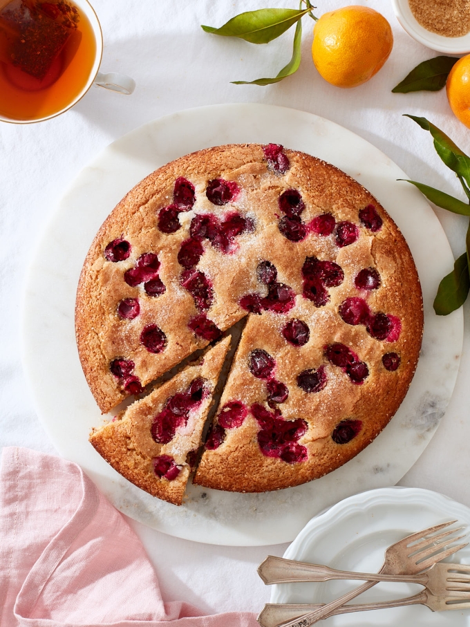 Citrus and Cranberry Buttermilk Cake | A winter cranberry cake on a white marble cake round that's just been sliced. White porcelain dessert plates with vintage forks, pink linen, tea, and fresh tangerines.