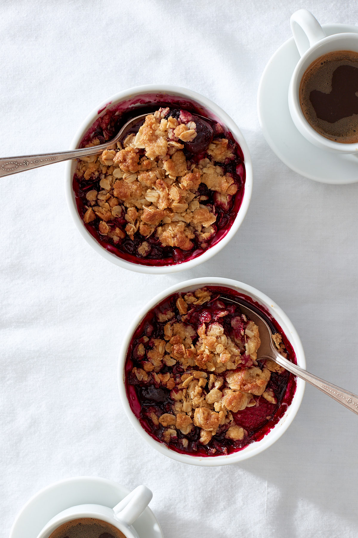 Two miniature mixed berry crumbles on a white linen background with little cups of espresso.