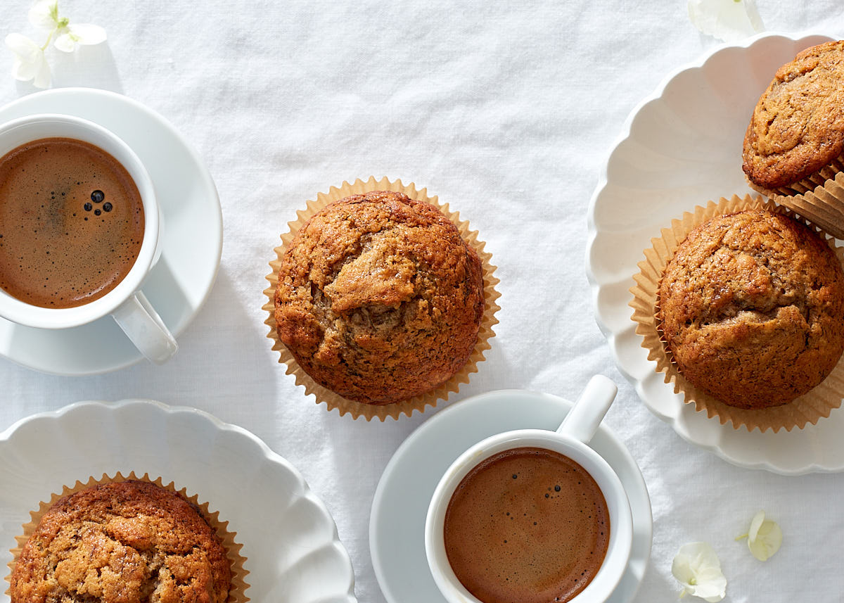 Banana muffins with two cups of espresso on fluted white china dessert plates and a white linen tablecloth.