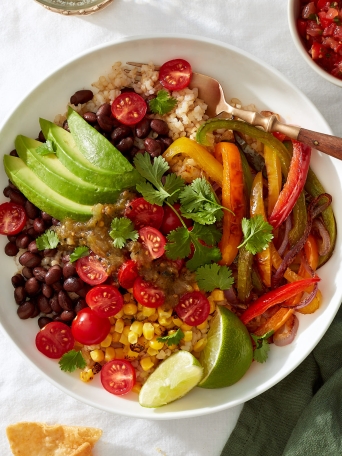 A vegetarian burrito bowl with rice, beans, fajita-style onions, peppers, and corn, avocado, salsa, cherry tomatoes, lime, and cilantro on a white linen tablecloth.