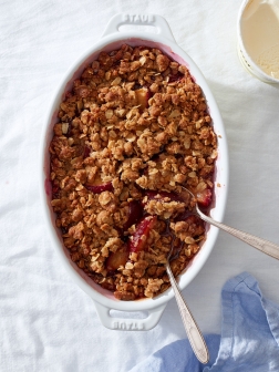 A plum crumble in a white baking dish with two vintage silver spoons.