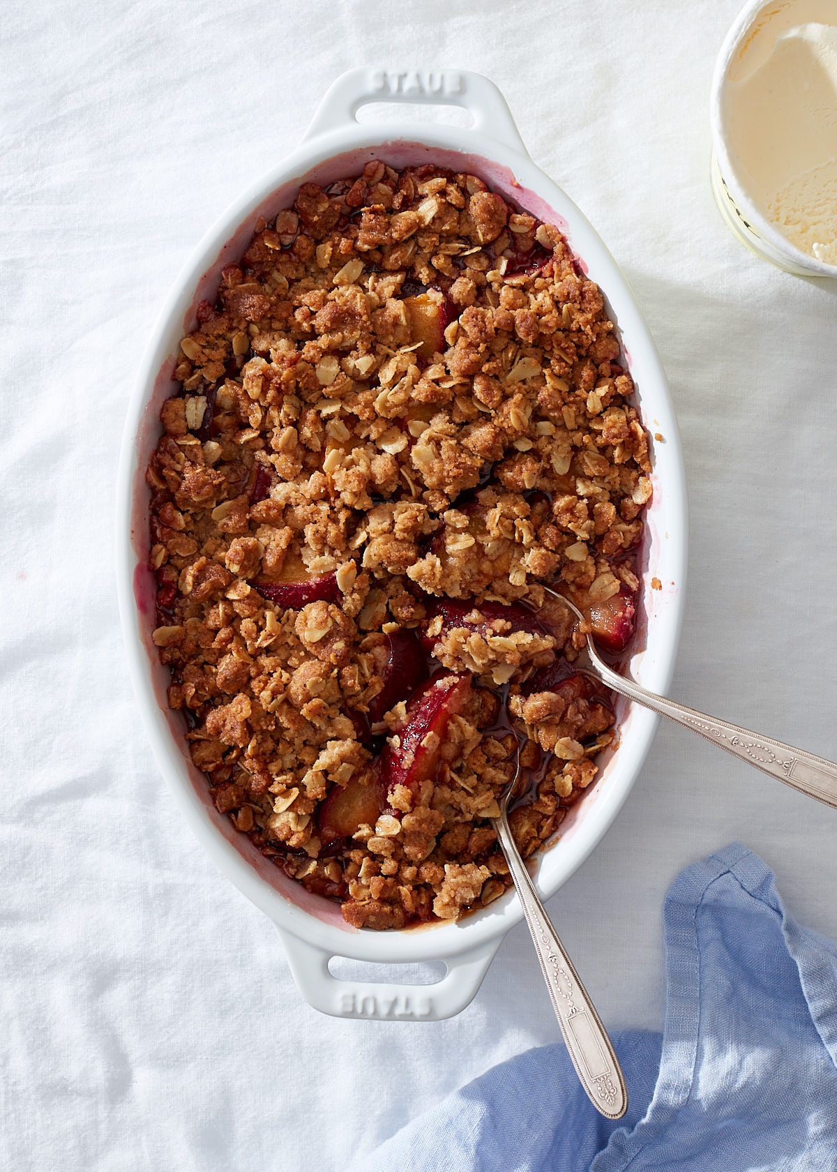 A plum crumble in a white baking dish with two vintage silver spoons.