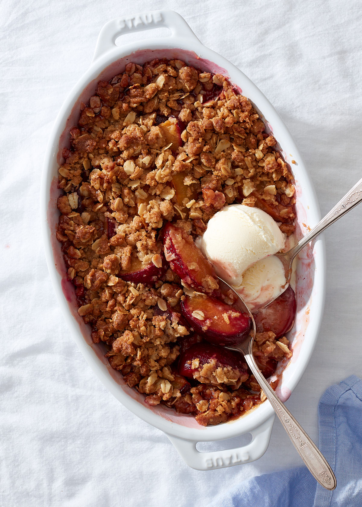 A plum crumble in a white baking dish with two vintage silver spoons and a scoop of vanilla ice cream.