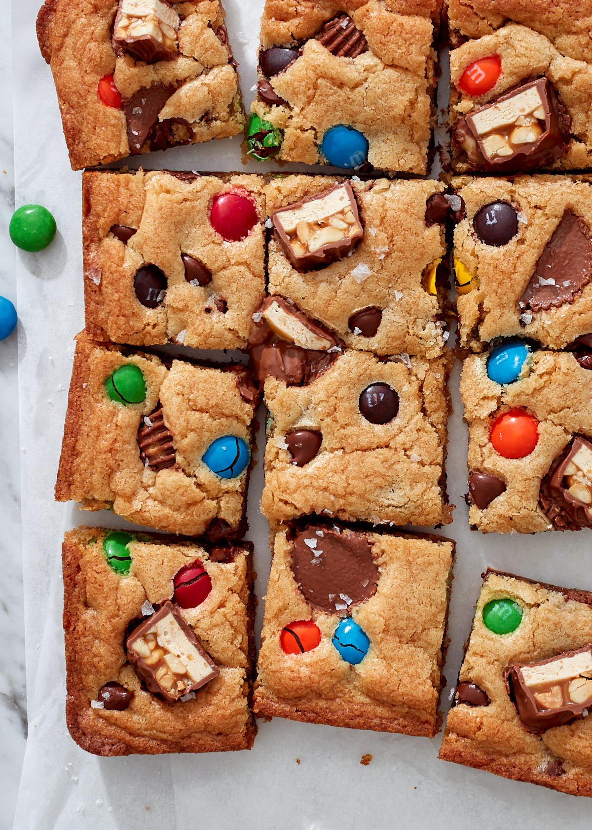 Candy bar blondies, with M&Ms, Snickers, and Reese's peanut butter cups on white parchment paper.