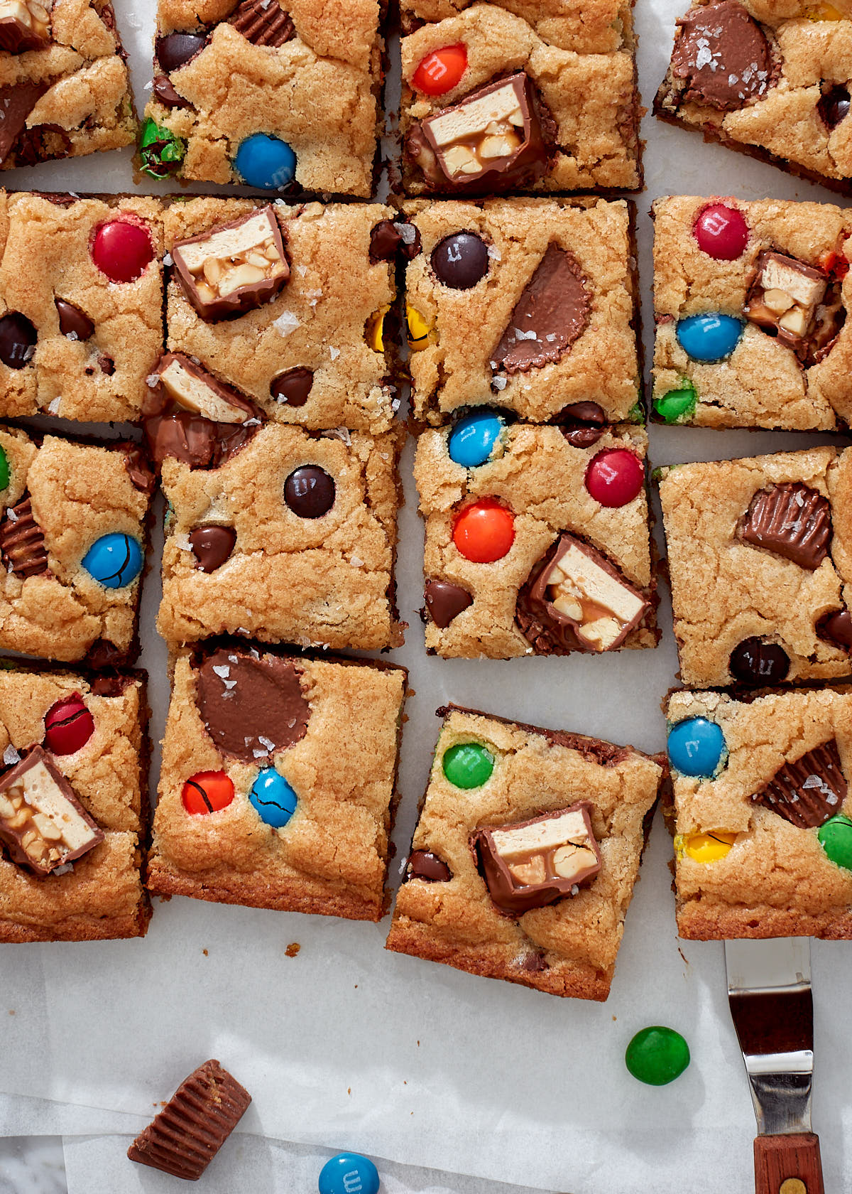 Candy bar blondies, with M&Ms, Snickers, and Reese's peanut butter cups on white parchment paper.