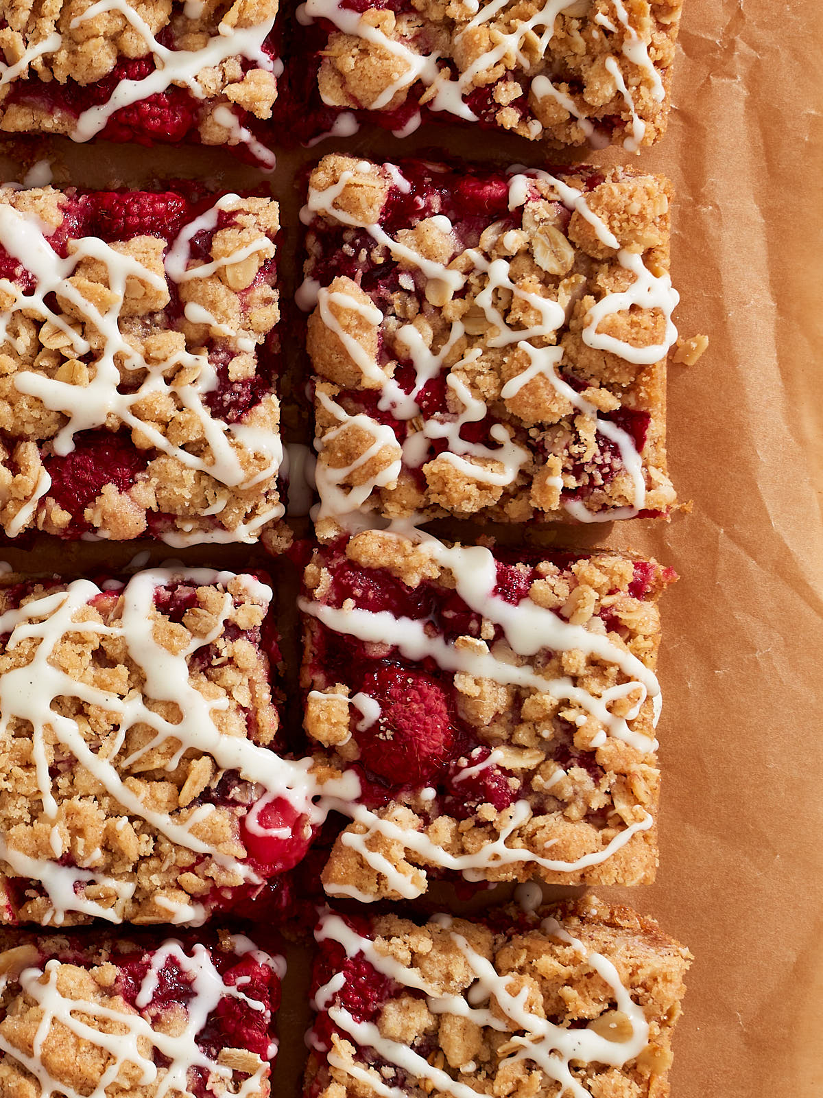 Rows of sliced raspberry and cranberry crumb bars drizzled with vanilla orange glaze on parchment paper.