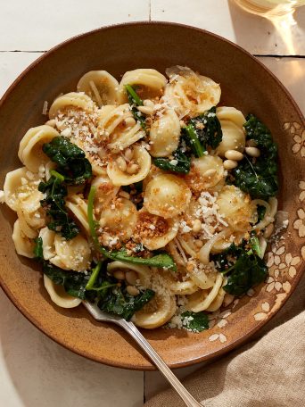 A bowl full of orecchiette pasta topped with wilted spinach, kale, toasted pine nuts, grated parmesan, and breadcrumbs.