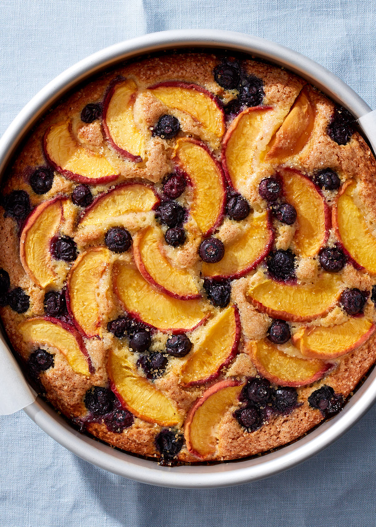 An everyday blueberry peach cake fresh from the oven in a silver baking pan on a light blue linen tablecloth.