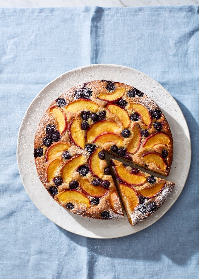 An blueberry peach cake on a white ceramic cake plate, with one slice cut, ready to be served. The background is a light blue linen tablecloth.