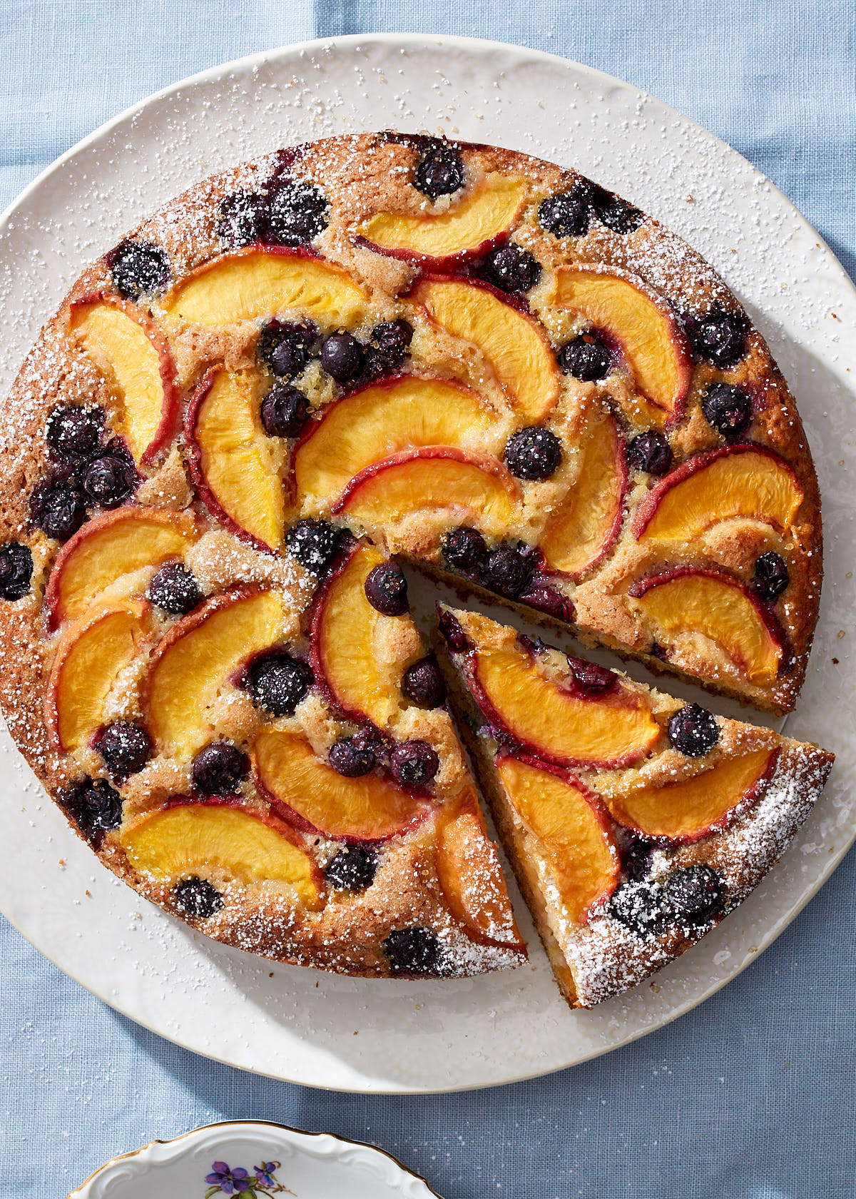 An blueberry peach cake on a white ceramic cake plate, with one slice cut, ready to be served. The background is a light blue linen tablecloth.